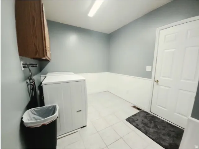 Laundry area featuring a wood-finish wall cabinet, light gray walls with white wainscoting, and white tiled flooring