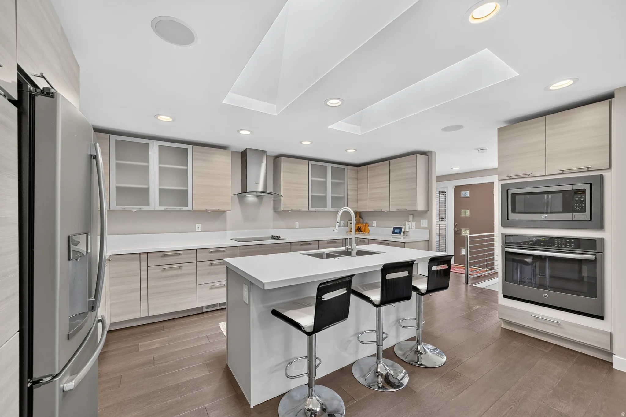 Spacious kitchen featuring an island with integrated sink, stainless steel appliances, light wood-finish cabinetry, recessed ceiling lighting, and a skylight