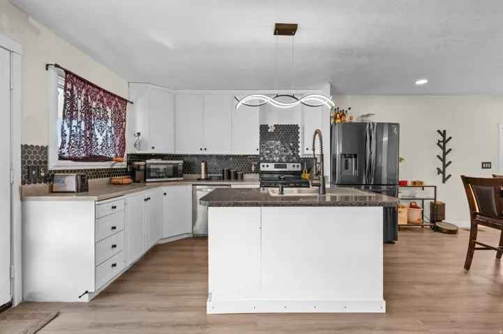 Kitchen featuring white cabinetry, a central island with a sink, stainless steel appliances, dark hexagonal tile backsplash, and wood-finish flooring
