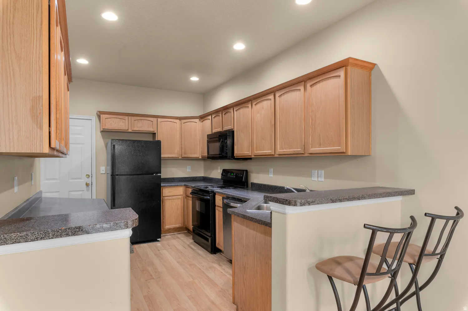 Spacious kitchen featuring wood cabinetry, dark countertops, full suite of black appliances, wood-finish flooring, and recessed lighting