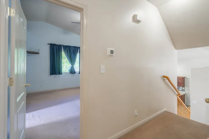 Carpeted hallway featuring a wooden handrail and banister, leading to a room with vaulted ceilings and a window