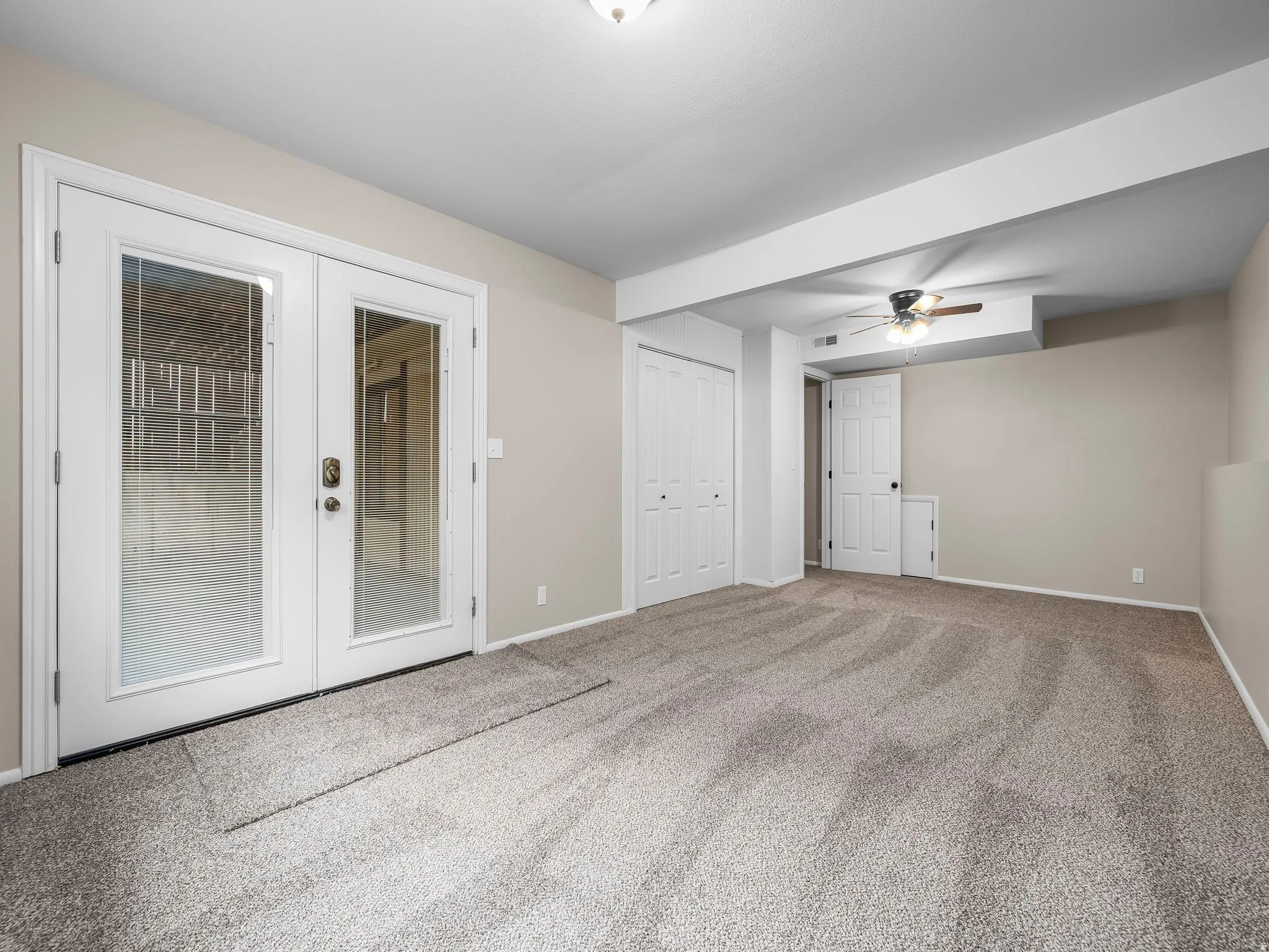 Carpeted room featuring white-framed glass paneled double doors, neutral painted walls, white bi-fold closet doors, a white interior door, and a ceiling fan with integrated lighting
