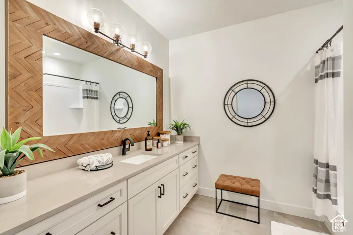 Bathroom featuring a long wood-framed mirror with a chevron pattern, a four-light vanity fixture with clear glass globes, a single undermount sink with a black faucet, white shaker-style cabinetry with black hardware, and light-toned countertop