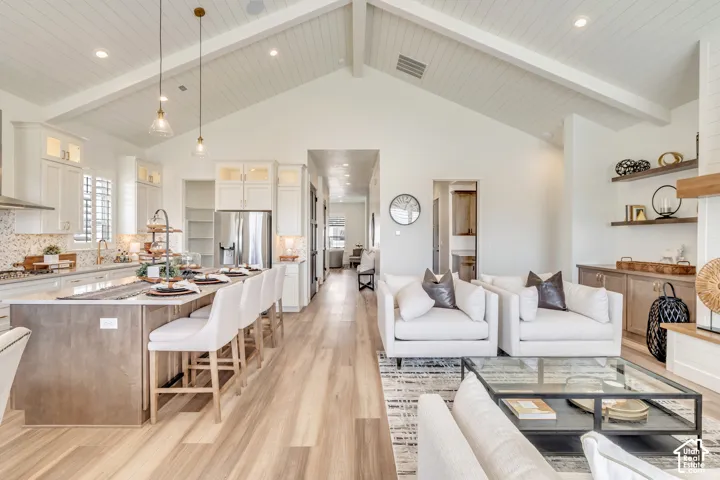 Vaulted ceiling with exposed beams, wood-finish flooring, built-in shelving, white cabinetry, and a large kitchen island with seating