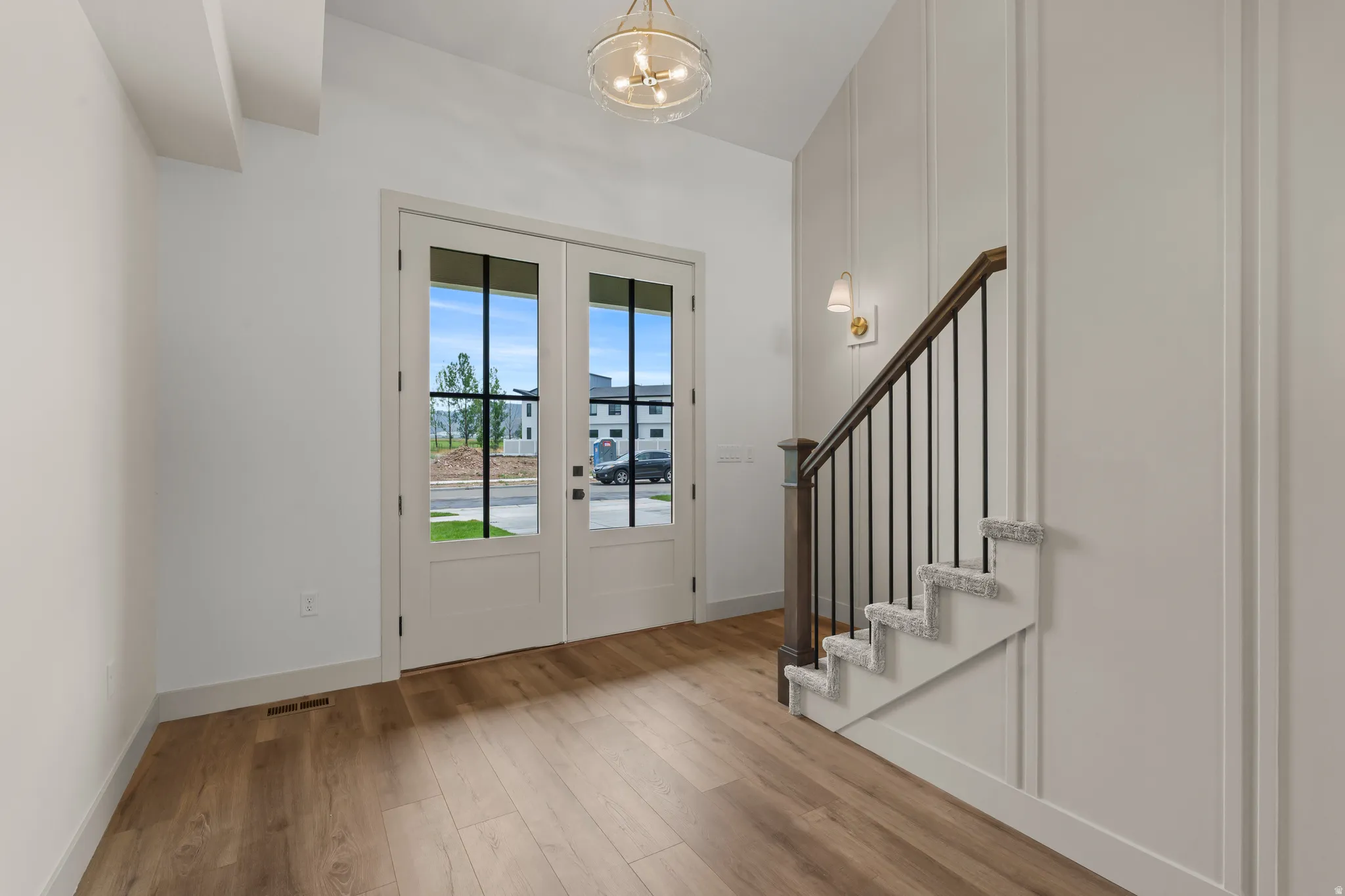 Entryway featuring double doors with divided lites, wood-finish flooring, a contemporary chandelier, a staircase with carpeting and wrought iron balusters, and wainscoting