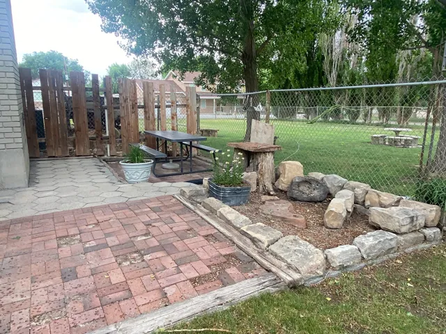 Outdoor patio featuring brick and paver surfaces, chain link fencing, a natural stone-lined garden bed, and mature trees