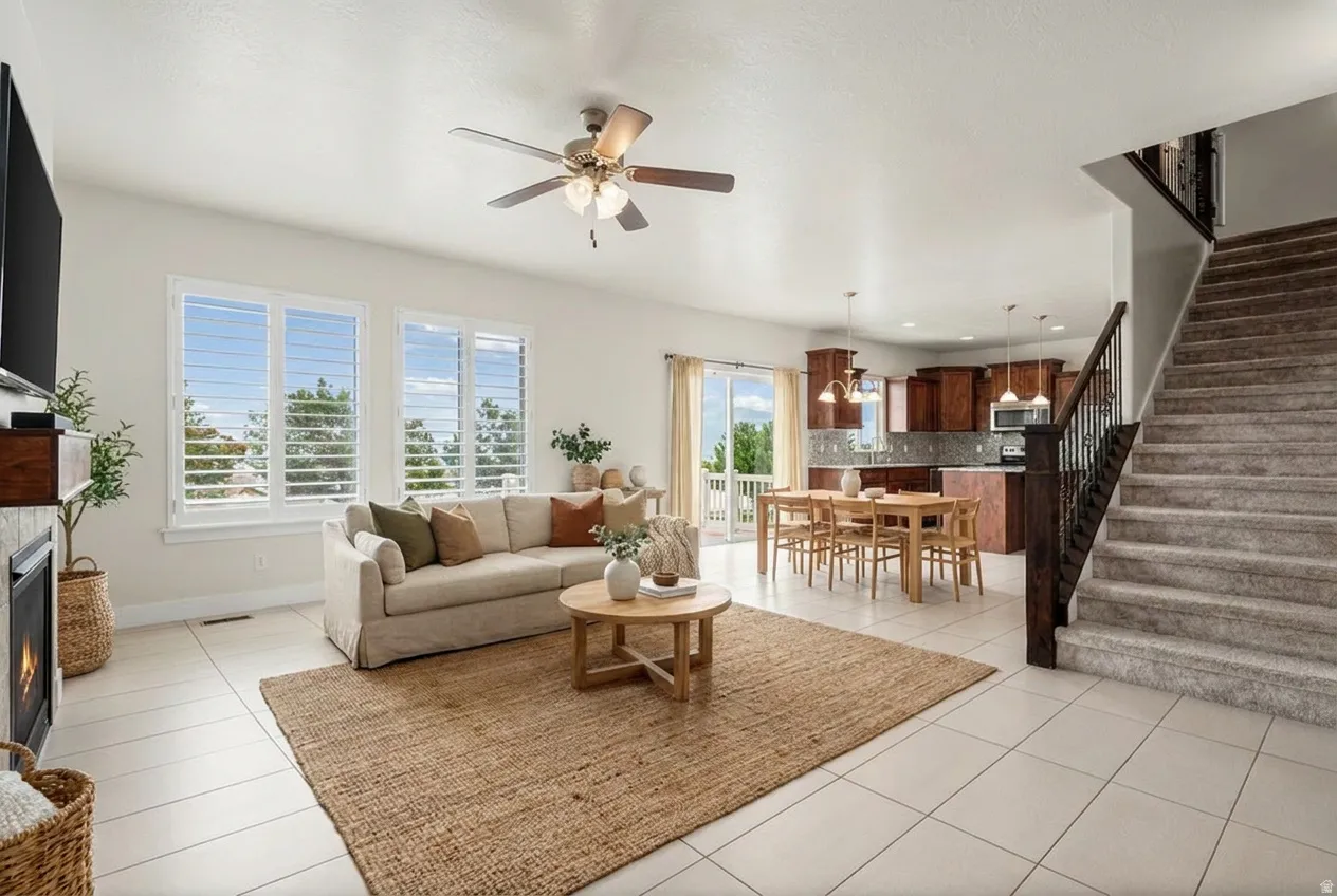 Open-concept living area with light-toned tile flooring, a ceiling fan, and multiple windows featuring plantation shutters. Virtually staged.