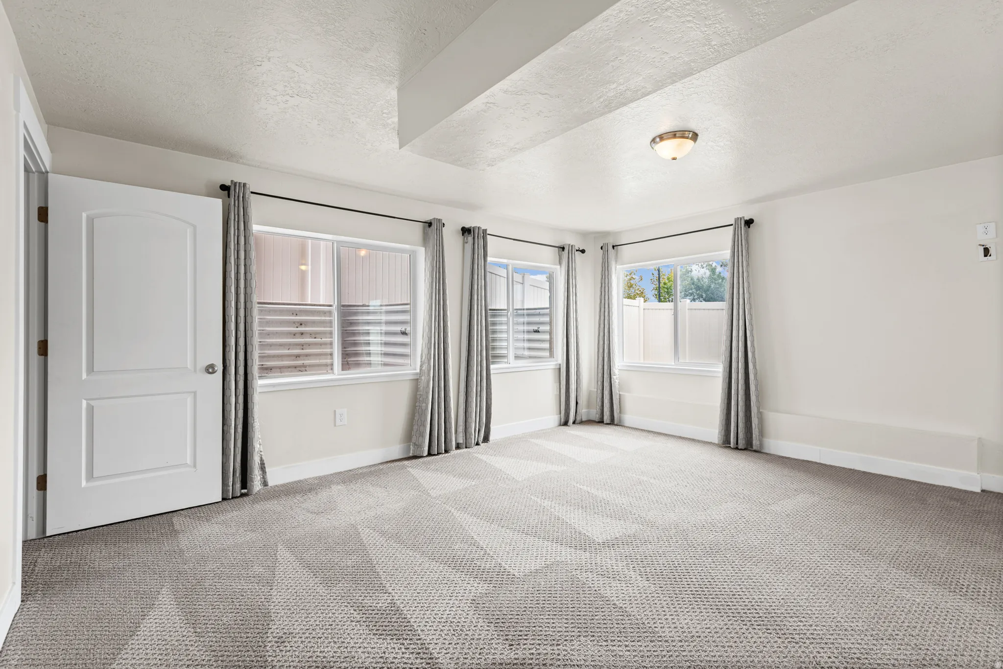 Spacious room featuring patterned gray carpet flooring, multiple windows with white trim, a semi-flush mount ceiling light fixture, a tray ceiling detail, and a paneled interior door