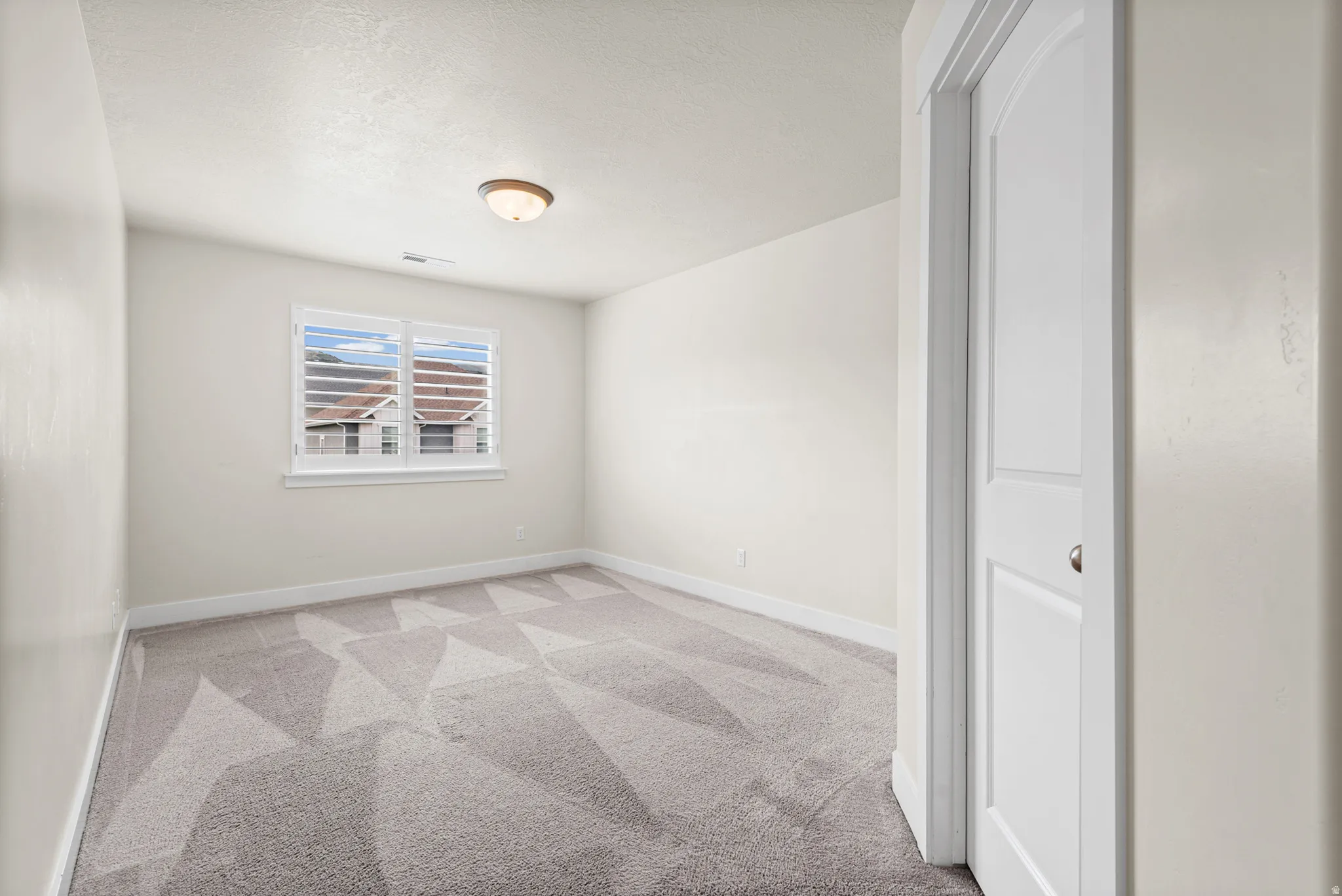 Neutral-toned room featuring a window with plantation shutters, a patterned gray carpet, white baseboards, and a flush-mount ceiling light