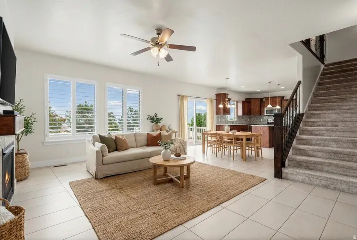 Open-concept living area with light-toned tile flooring, a ceiling fan, and multiple windows featuring plantation shutters. Virtually staged.