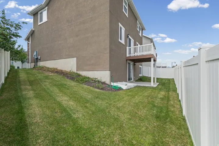 Expansive grassy yard featuring a stucco exterior, white vinyl fencing, a ground-level concrete patio, and a second-story balcony with white railings