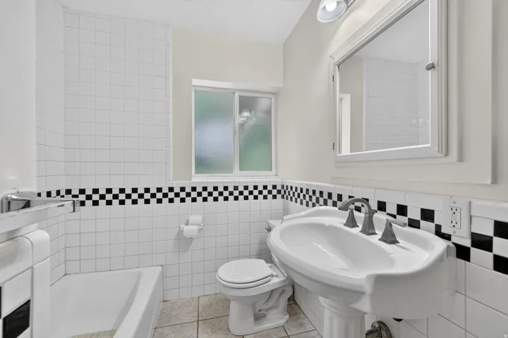 Bathroom featuring a pedestal sink, white tile walls with a black and white checkerboard accent, white toilet, and a frosted window