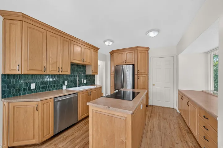 Kitchen featuring natural wood cabinetry, a striking green geometric tile backsplash, a center island with a built-in cooktop, stainless steel appliances, and wood-finish flooring
