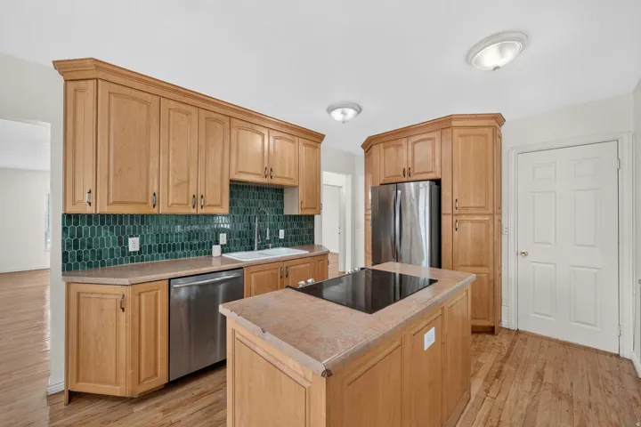 Kitchen featuring light wood cabinetry, a central island with an integrated cooktop, stainless steel appliances, and a green geometric tile backsplash