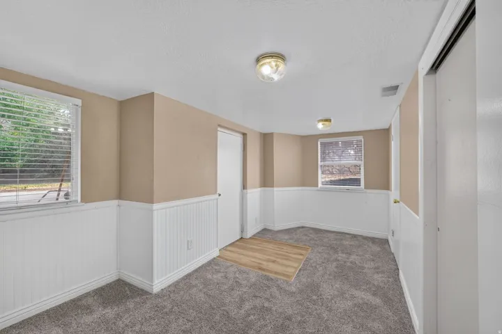 Inviting room featuring a window with horizontal blinds, neutral wall paint, white wainscoting, and gray textured carpeting with a wood-finish flooring insert