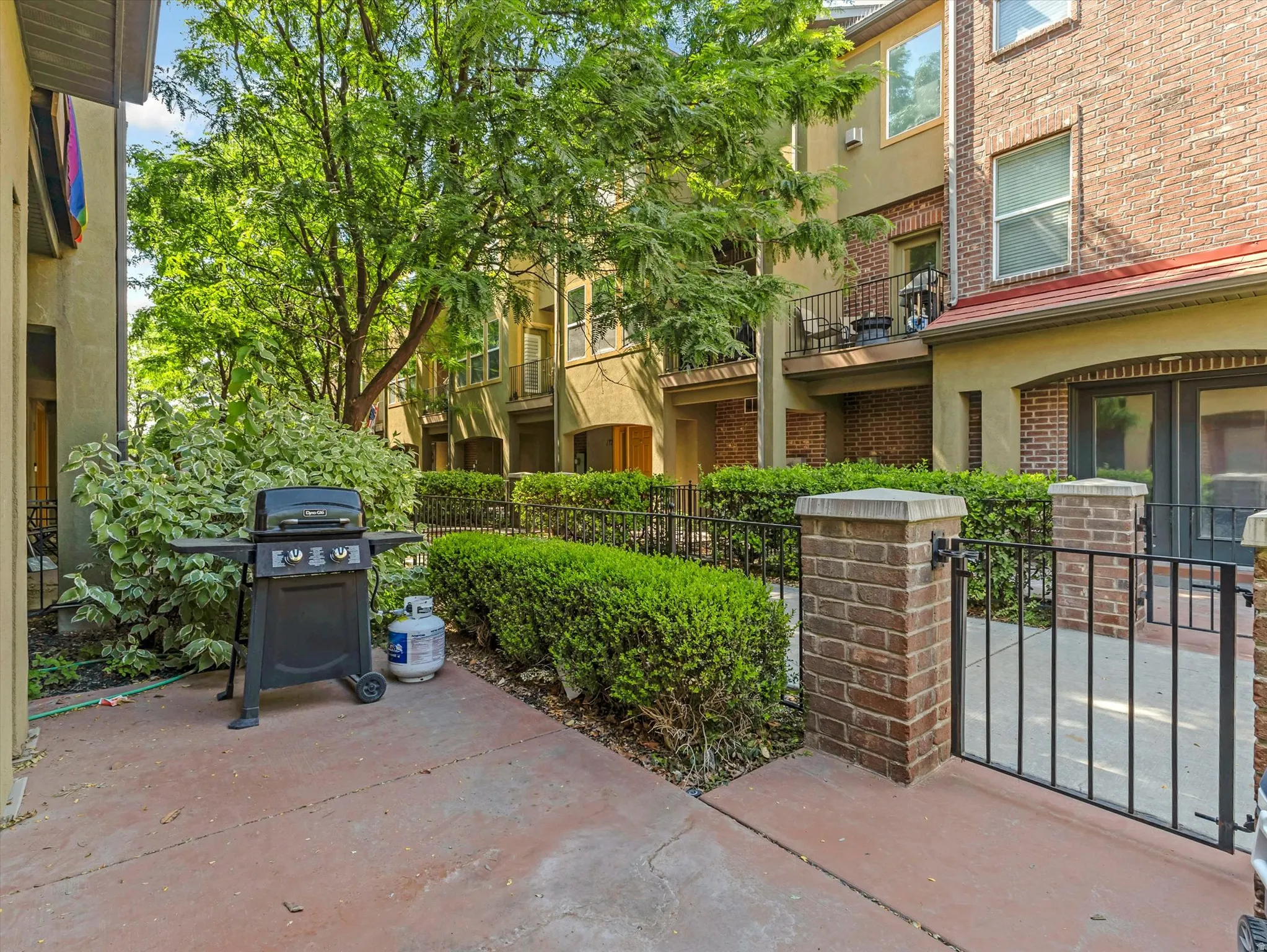 Private patio space featuring a concrete surface, mature landscaping, and a wrought iron gate with brick pillars