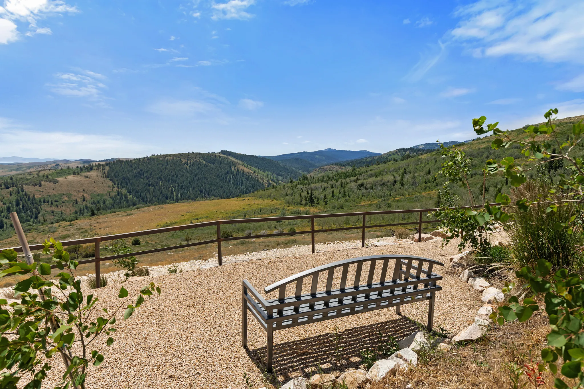 Outdoor gravel patio with a metal bench, framed by a wooden fence, overlooking a vast mountainous landscape with dense tree coverage and natural vegetation
