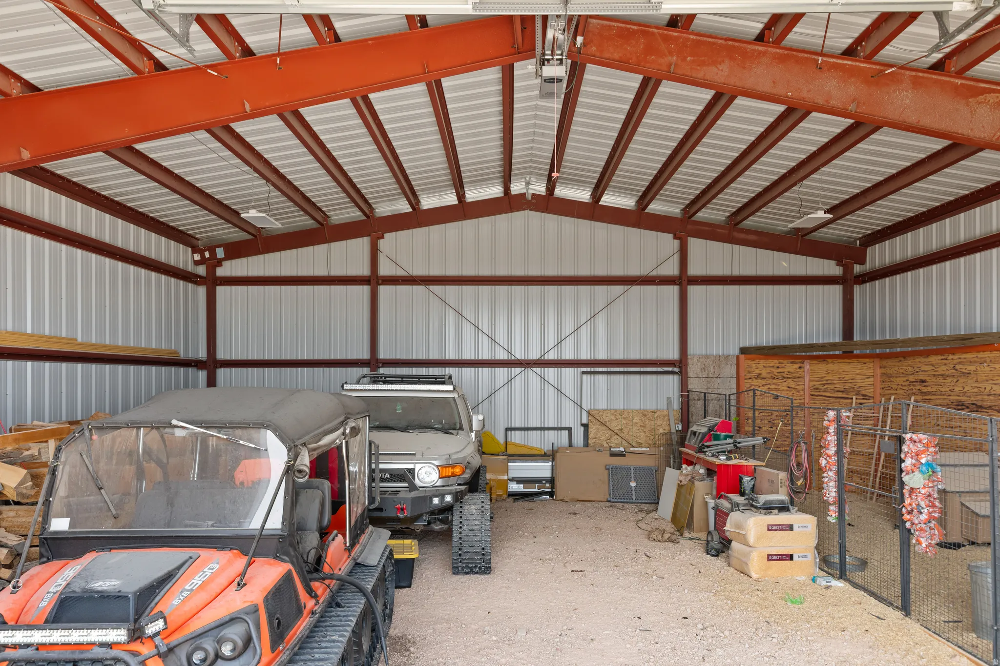 Spacious metal building featuring a vaulted ceiling with exposed red structural beams, corrugated metal walls, and a gravel floor