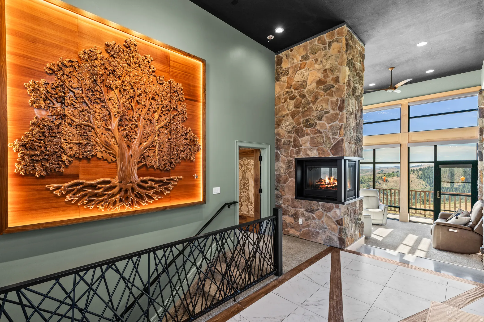 Two-story stone fireplace with a three-sided hearth, a custom wood-carved wall art installation with backlighting, and an open living space with expansive windows