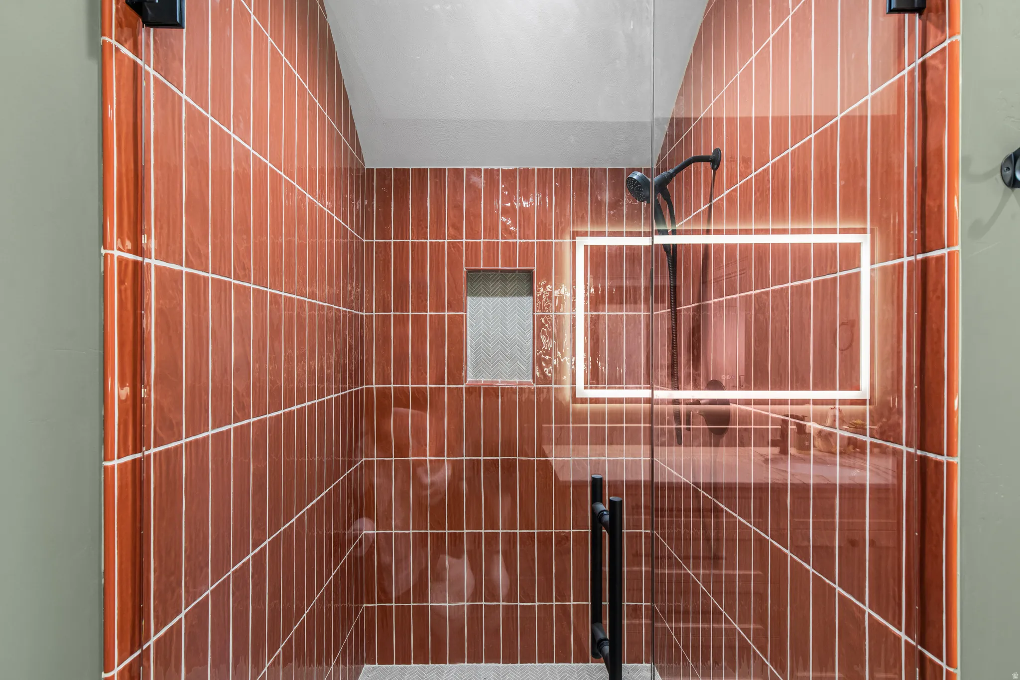 Glass-enclosed shower featuring vertically-stacked terracotta-hued wall tiles, black matte fixtures, and a recessed tiled niche