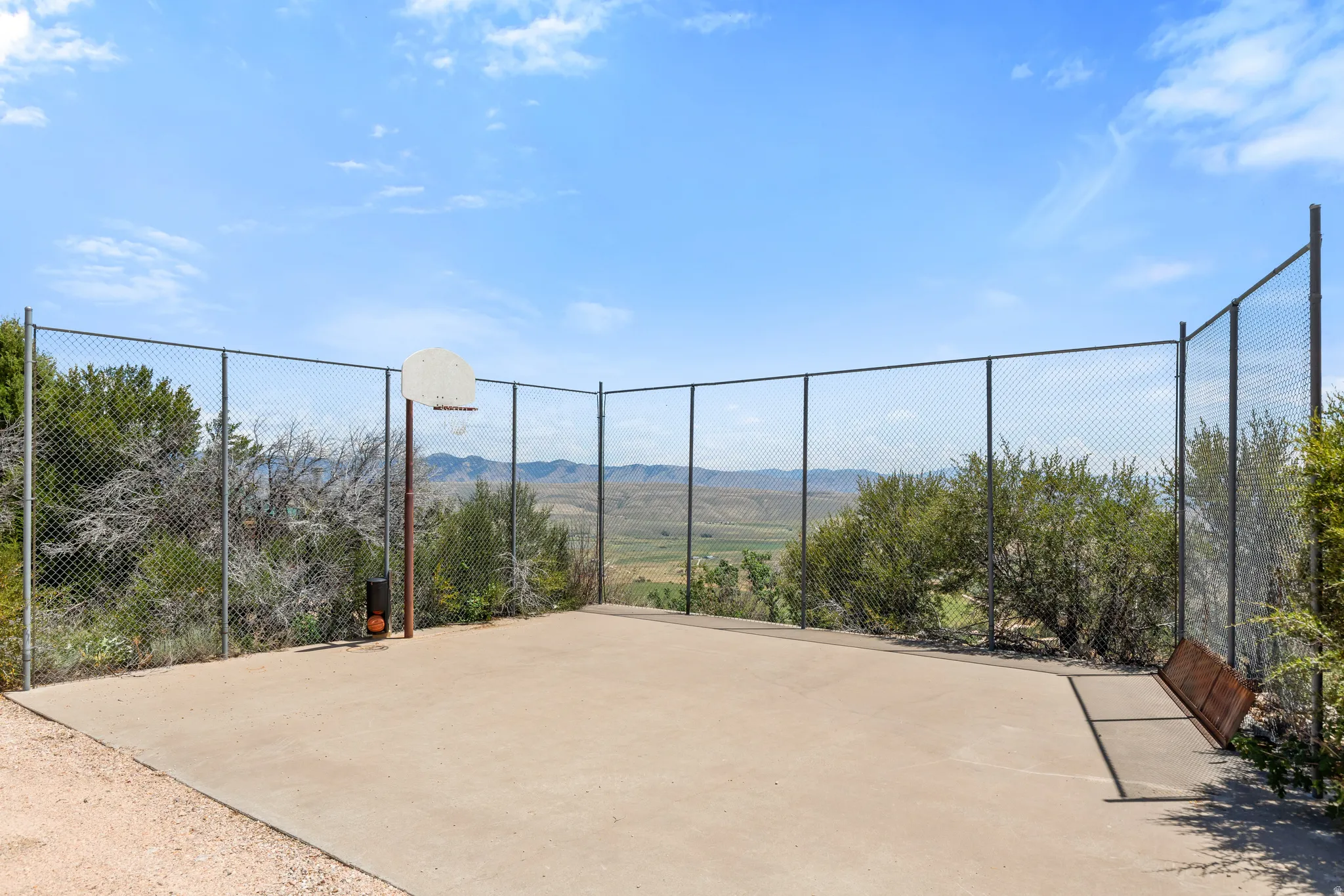Paved outdoor court featuring a basketball hoop with a metal backboard, surrounded by chain-link fencing