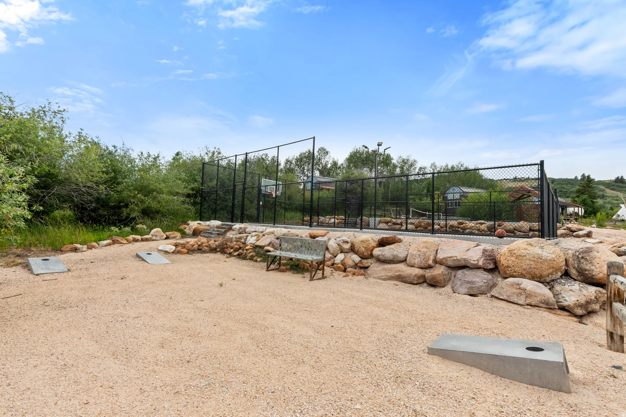 Outdoor recreational area featuring a fenced court with a basketball hoop, a gravel activity space, and a natural stone retaining wall