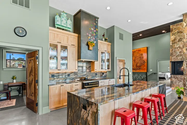Open-concept kitchen with a large island featuring a stone countertop, light wood cabinetry, a built-in range, and a decorative range hood