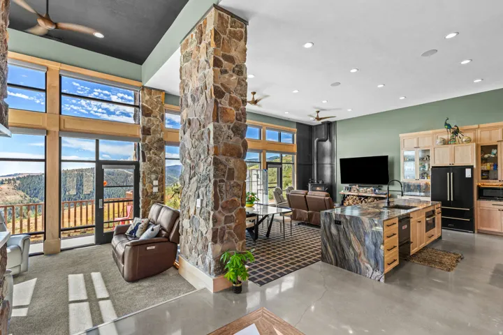 Open-concept living space featuring expansive floor-to-ceiling windows, stone-clad architectural pillars, a kitchen island with a waterfall countertop, light wood cabinetry, and polished concrete flooring