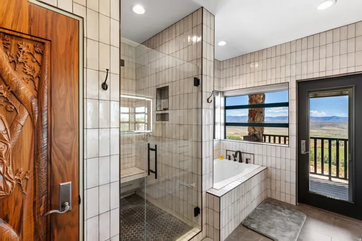 Bathroom featuring a clear glass shower enclosure, a built-in tub, white vertical tile accents, a private exterior balcony, and a carved wood door
