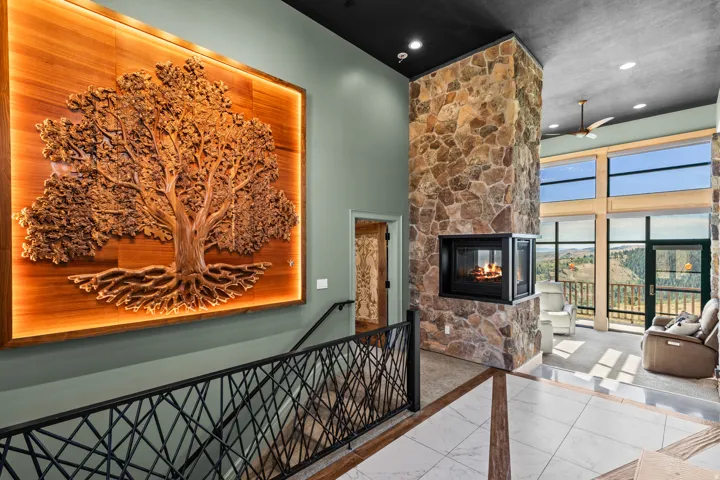 Two-story stone fireplace with a three-sided hearth, a custom wood-carved wall art installation with backlighting, and an open living space with expansive windows