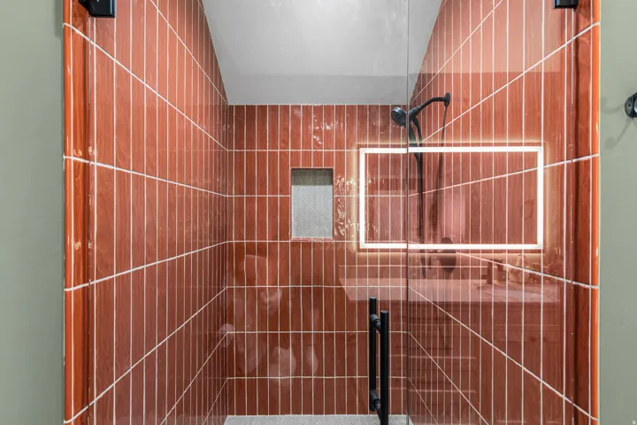 Glass-enclosed shower featuring vertically-stacked terracotta-hued wall tiles, black matte fixtures, and a recessed tiled niche