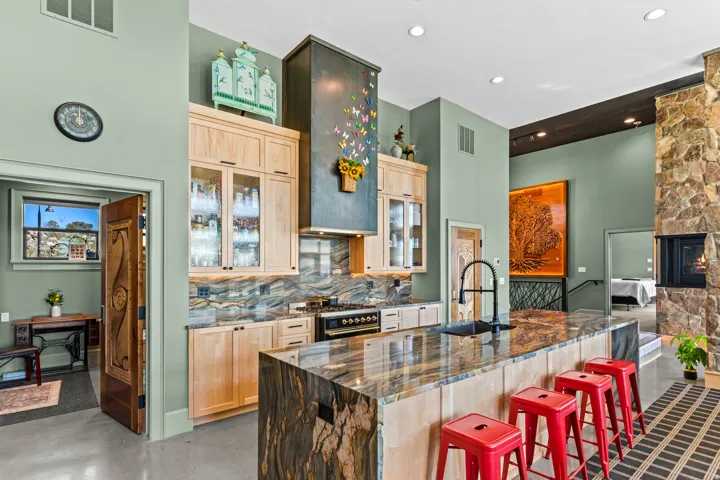 Open-concept kitchen with a large island featuring a stone countertop, light wood cabinetry, a built-in range, and a decorative range hood