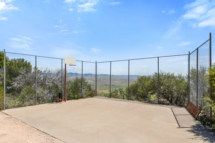 Paved outdoor court featuring a basketball hoop with a metal backboard, surrounded by chain-link fencing