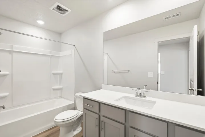Full bathroom featuring a white tub and shower combination with built-in shelving, a toilet, a vanity with a white countertop and undermount sink, a chrome faucet, and gray cabinetry