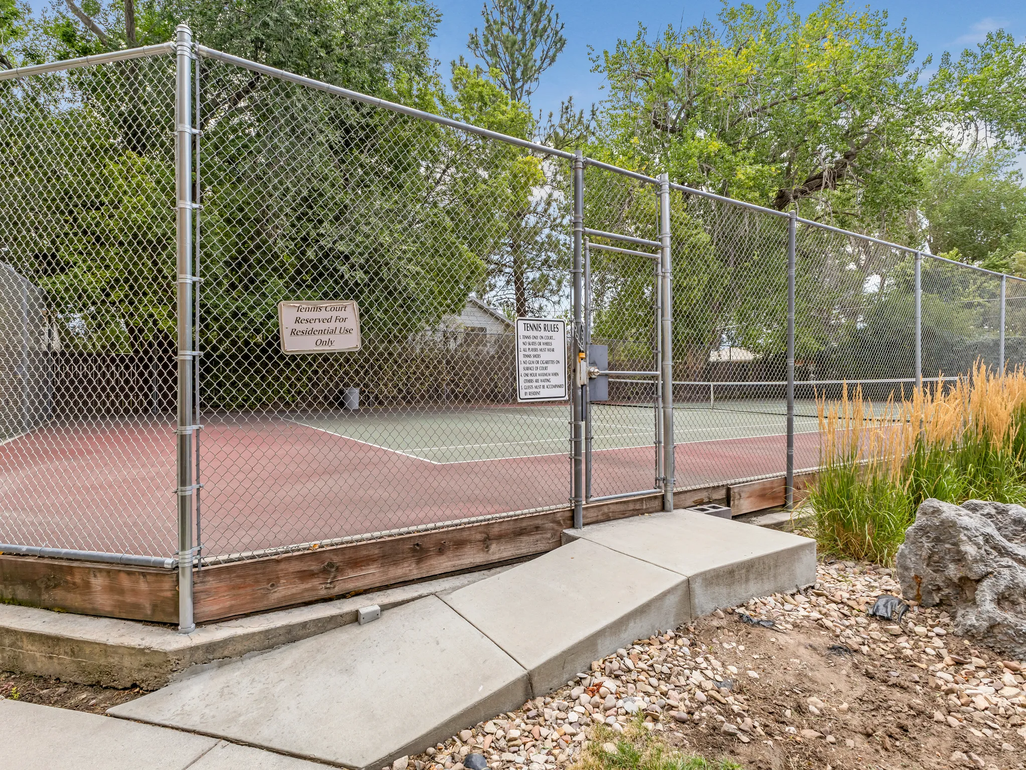 Fenced tennis court featuring a red and green playing surface, chain-link enclosure, and a concrete access ramp