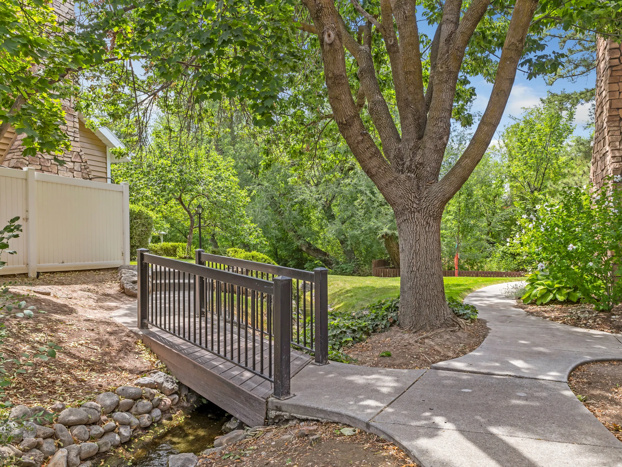 Landscaped grounds featuring a dark wood-finish footbridge over a small creek, surrounded by mature trees and a manicured lawn