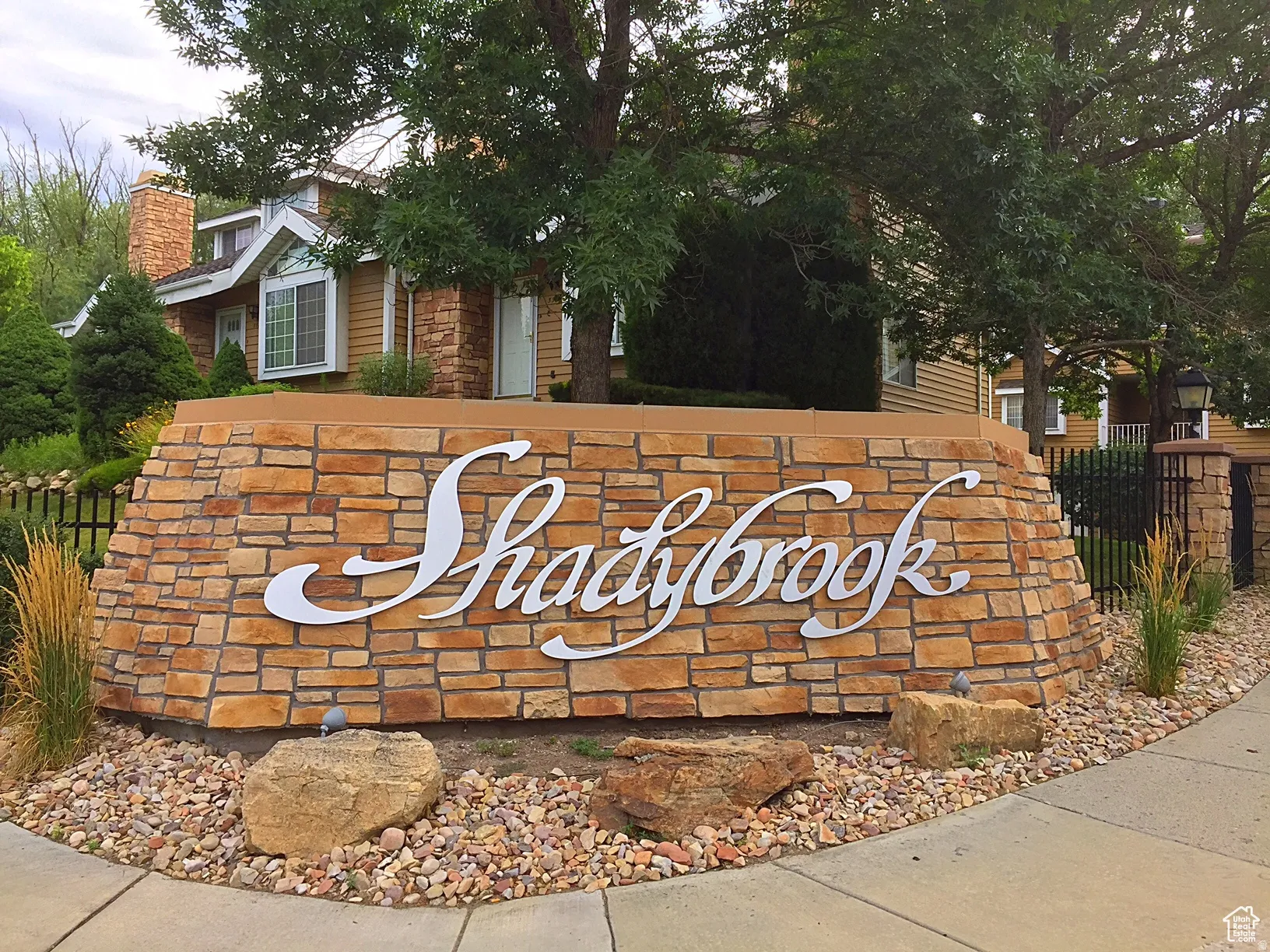 Stone veneer community sign featuring dimensional lettering, surrounded by river rock landscaping with mature trees and shrubbery
