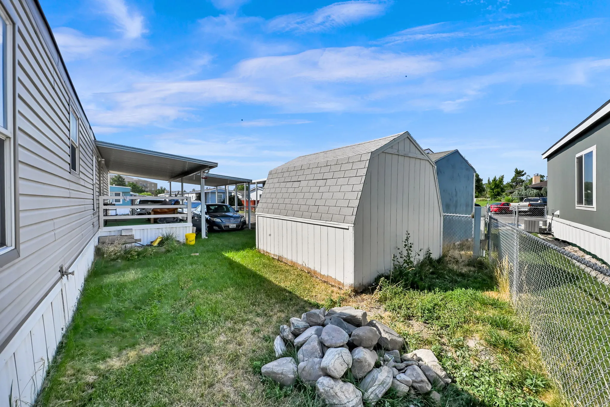 Grassed side yard featuring a storage shed with gambrel roof, chain link fencing, and a covered carport structure