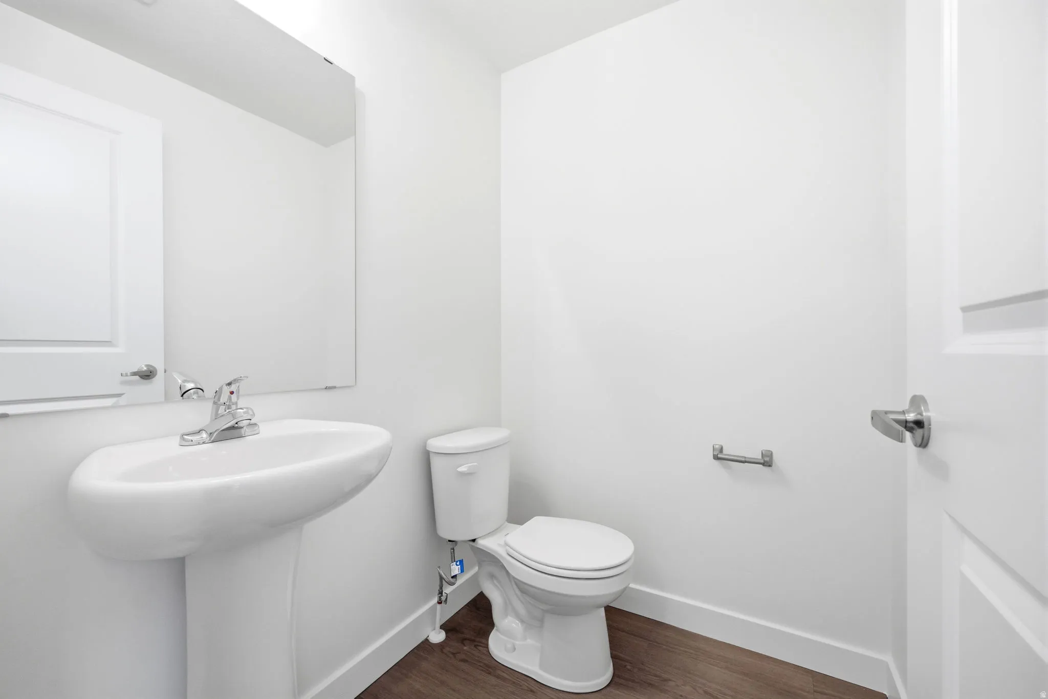 Pedestal sink with chrome faucet, wall-mounted mirror, toilet, wood-finish flooring, and white interior door