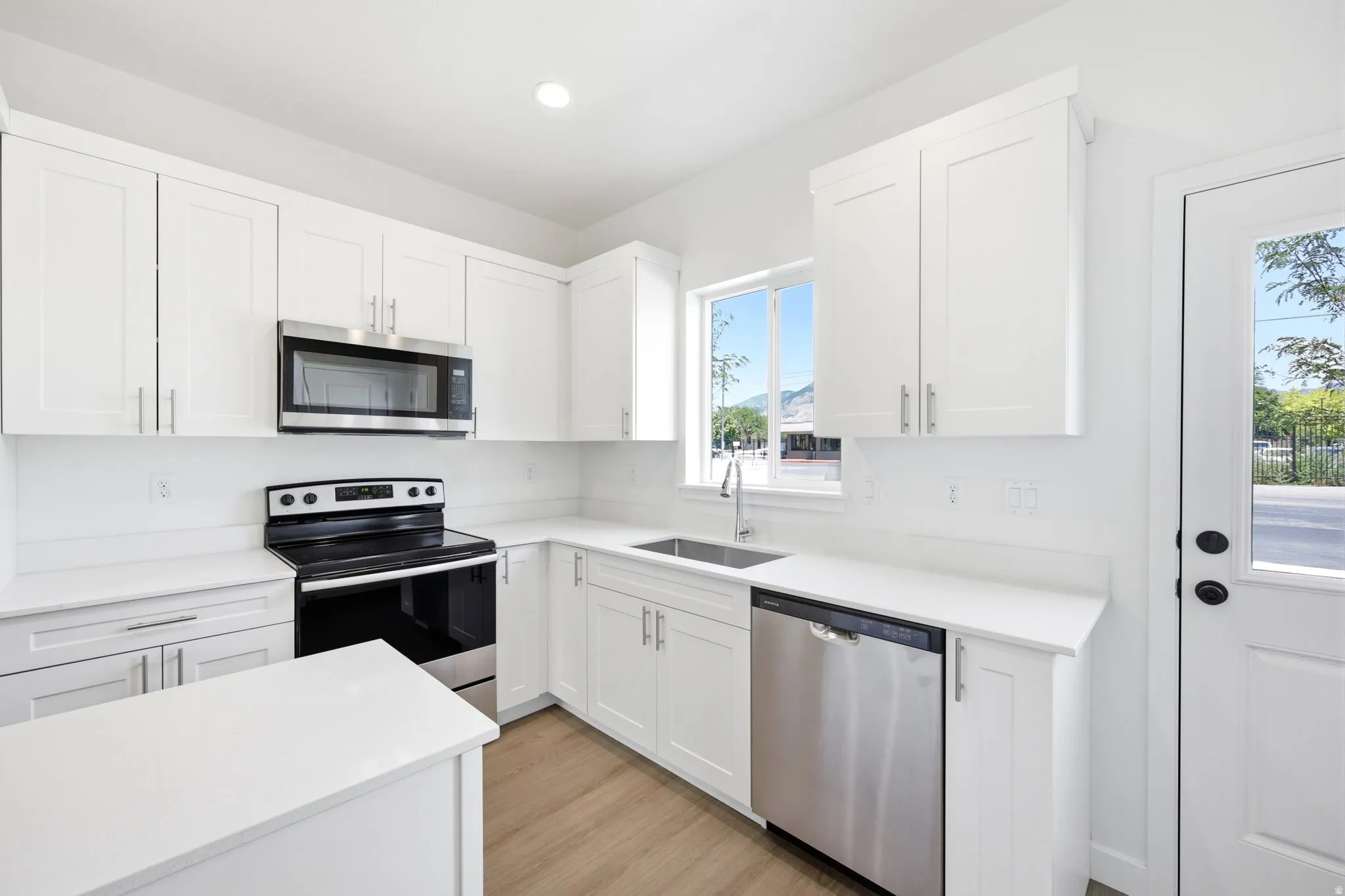 Modern kitchen featuring white cabinetry, stainless steel appliances, light-toned countertops, wood-finish flooring, and a bright window