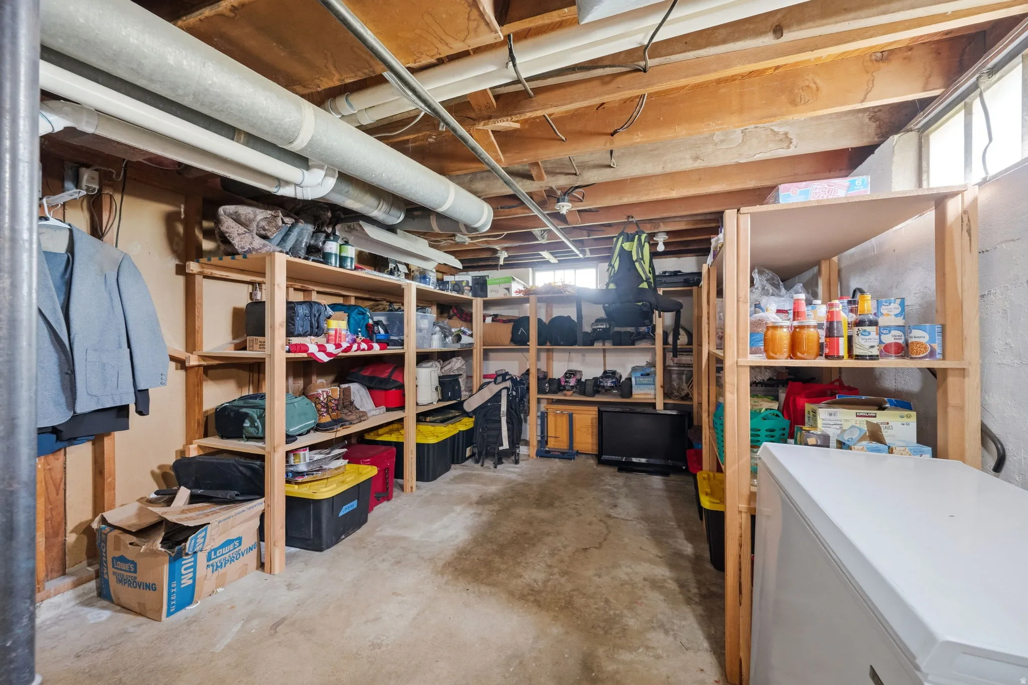 Basement storage area featuring extensive wood shelving, exposed ceiling joists, concrete flooring, and high-set windows