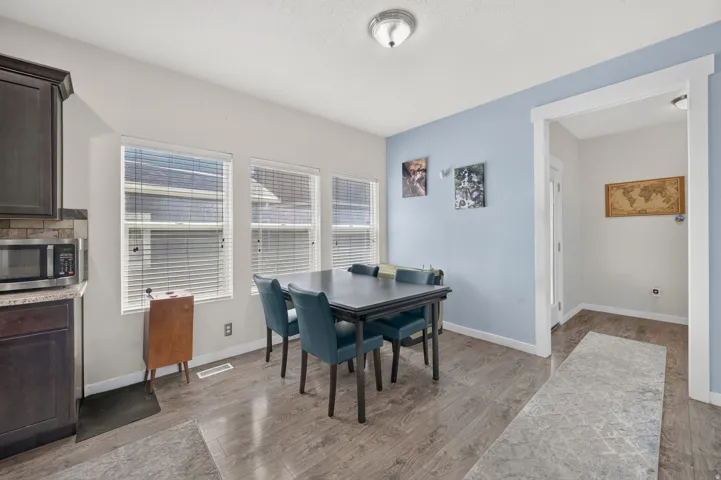 Dining area with wood-finish flooring, three windows with blinds, a light blue accent wall, and a built-in kitchen cabinet with stone-look countertops and a tile backsplash