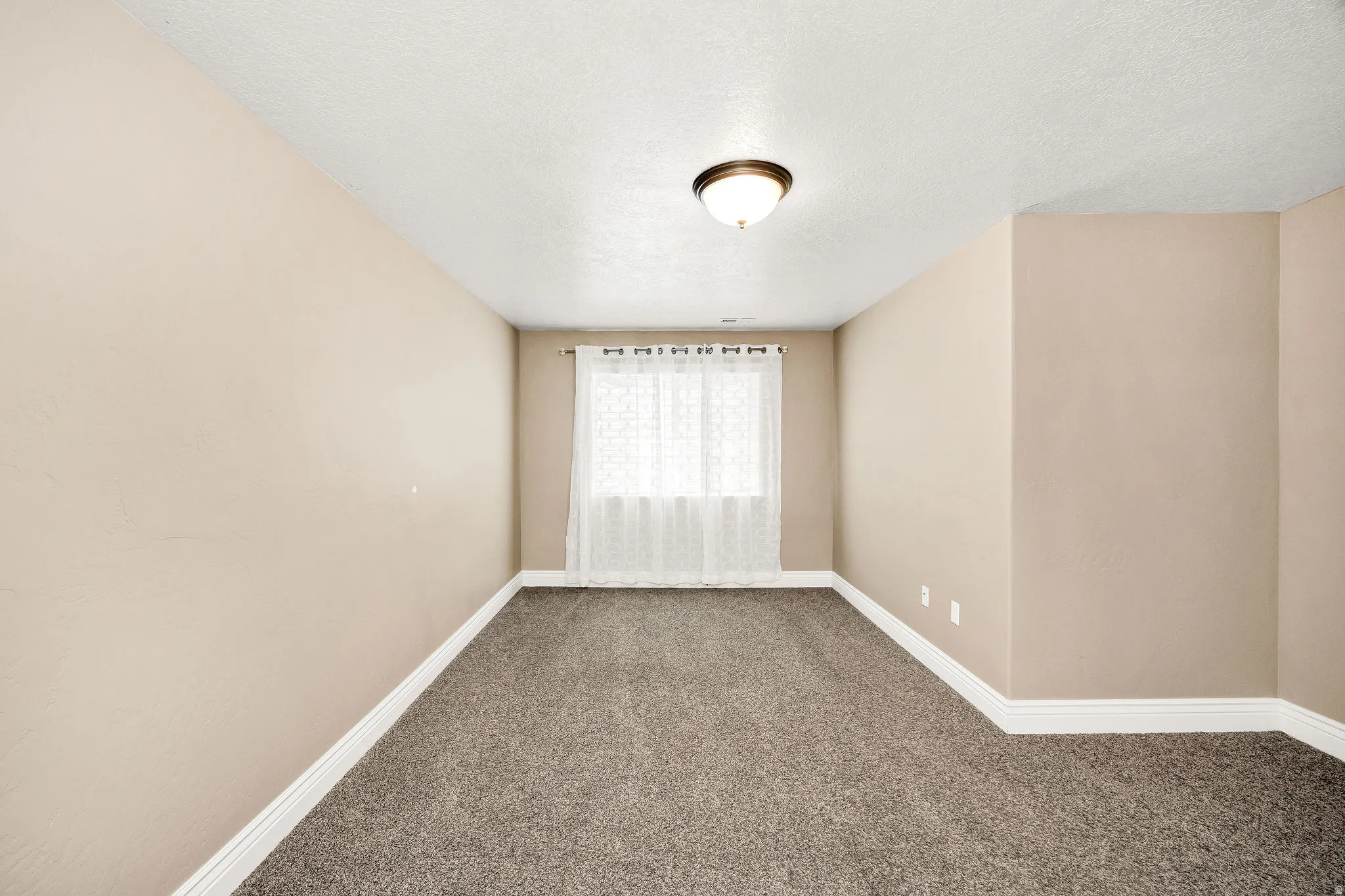 Carpeted room featuring a window with white sheer curtains, neutral-toned walls, white baseboards, and a ceiling-mounted light fixture
