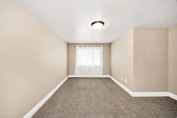 Carpeted room featuring a window with white sheer curtains, neutral-toned walls, white baseboards, and a ceiling-mounted light fixture