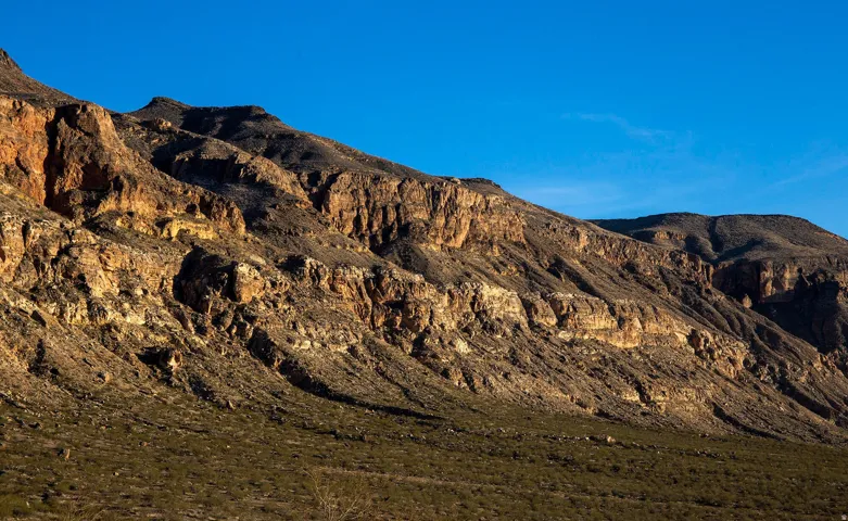Mountainous terrain featuring layered rock formations and arid vegetation under a clear sky