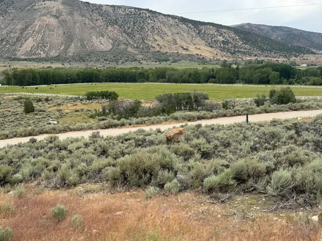 Mountainous landscape featuring a broad green field, natural foliage, and a rustic dirt road