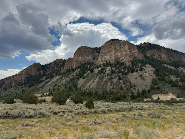 Mountain landscape featuring rocky cliffs, forested slopes, and high desert shrubland