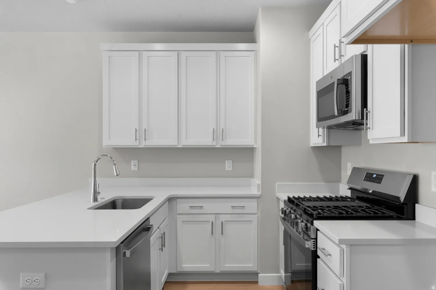 White shaker cabinetry, stainless steel appliances, corner sink, light-toned countertops, and wood-finish flooring