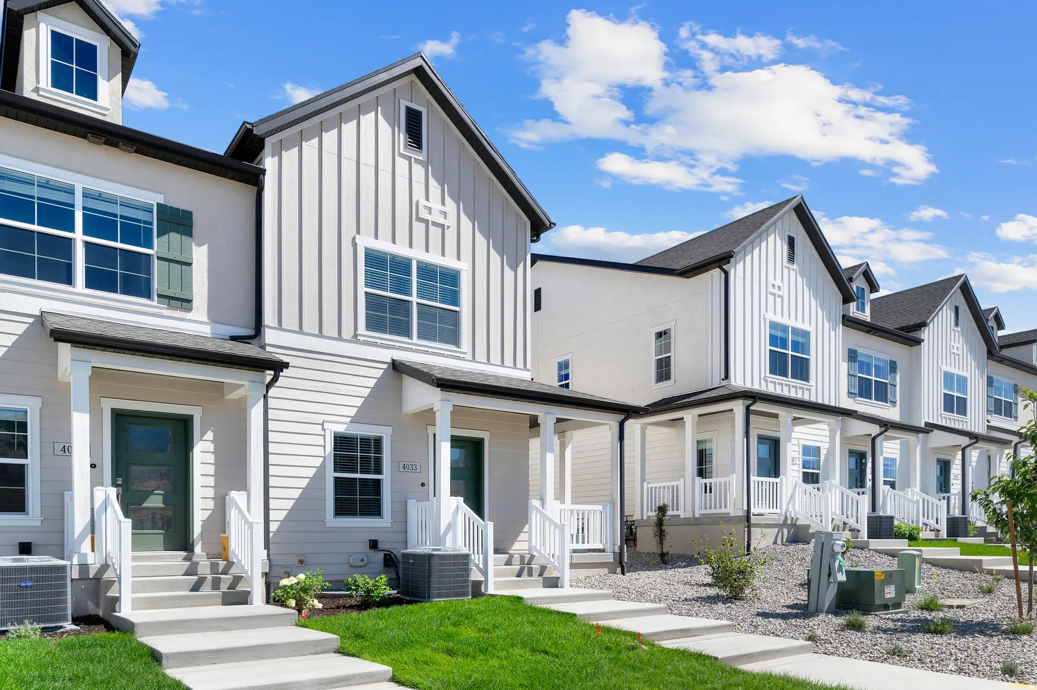 Row of townhomes featuring board-and-batten and horizontal siding, covered front porches with white railings, dormer windows, and gabled rooflines