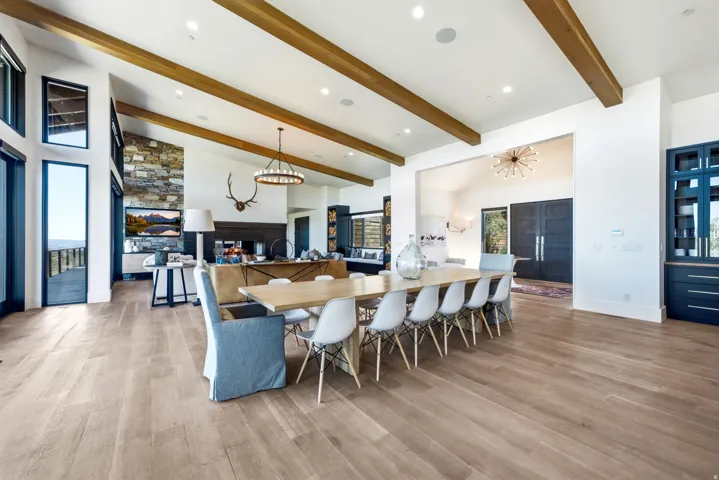 Spacious interior featuring wood-finish flooring, vaulted ceilings with exposed wood beams, a stone accent wall, large floor-to-ceiling windows, and a built-in dark wood cabinetry with glass fronts